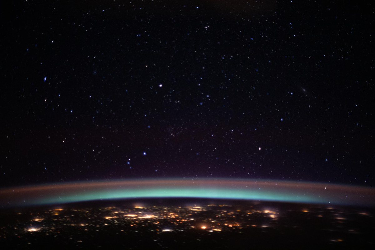 Many stars above a glowing curved band of air. Below, Earth is dark with clusters of orange city lights.