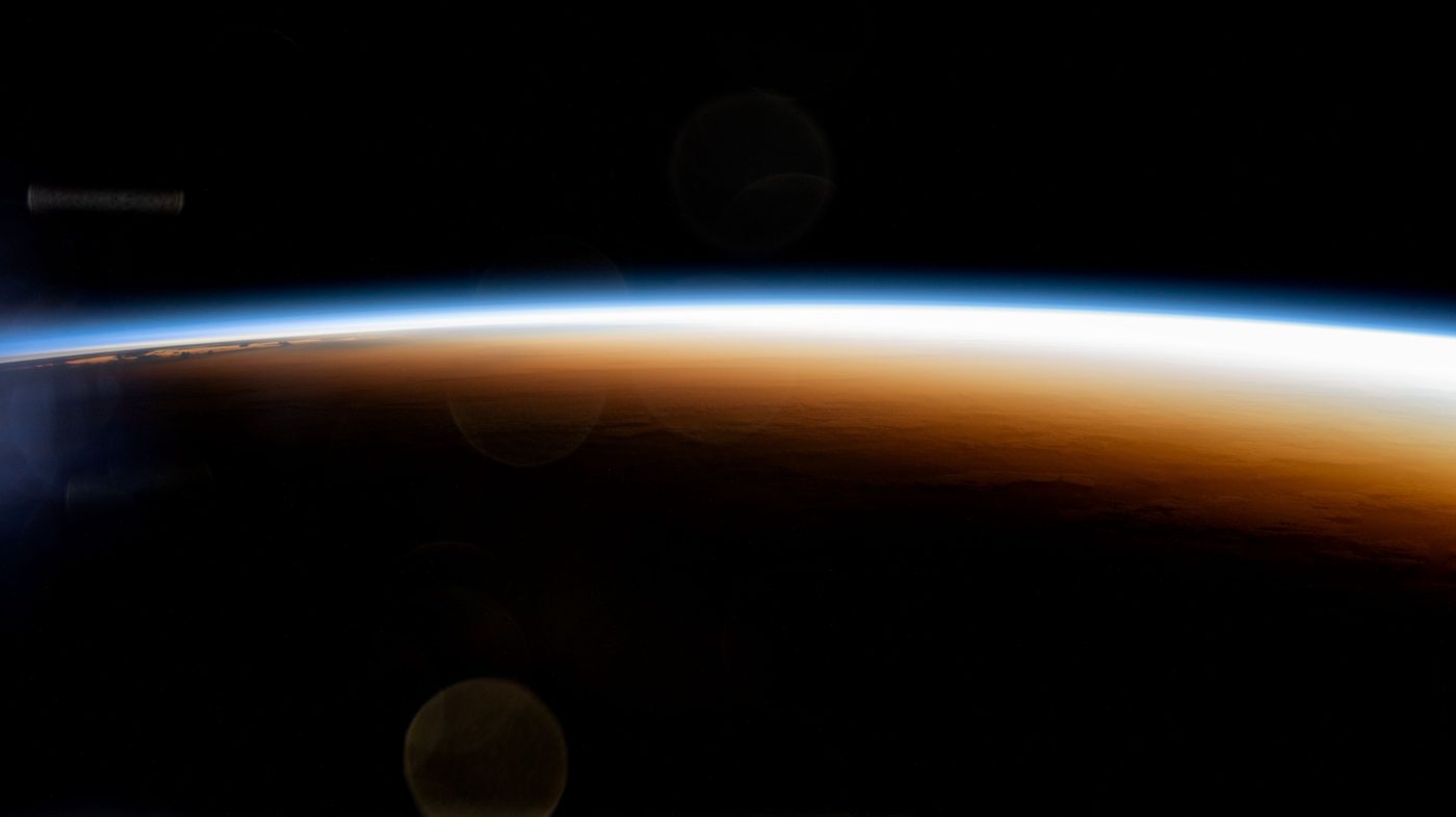 A thin glowing band of sunrise curves along the edge of Earth, with dark night below it and black space above.
