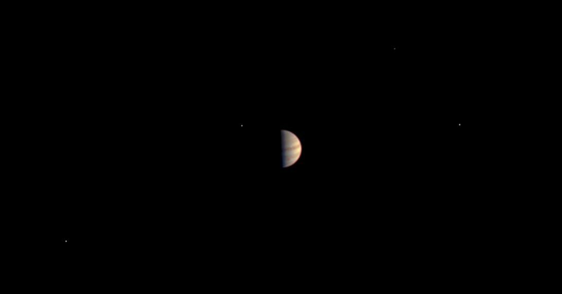 Jupiter as a half-lit striped disk in a black sky, with four tiny dots of light spread around it.